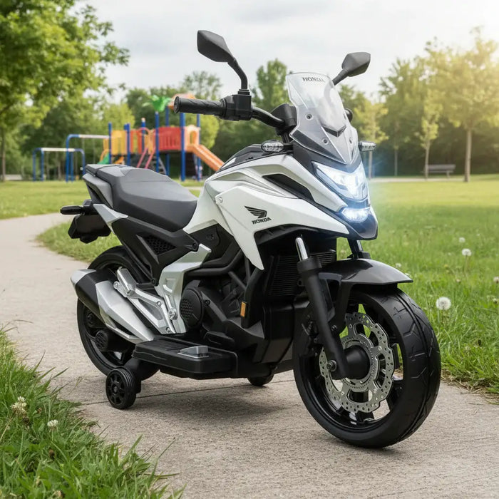White Honda electric toy motorcycle for kids with training wheels on park pathway near playground