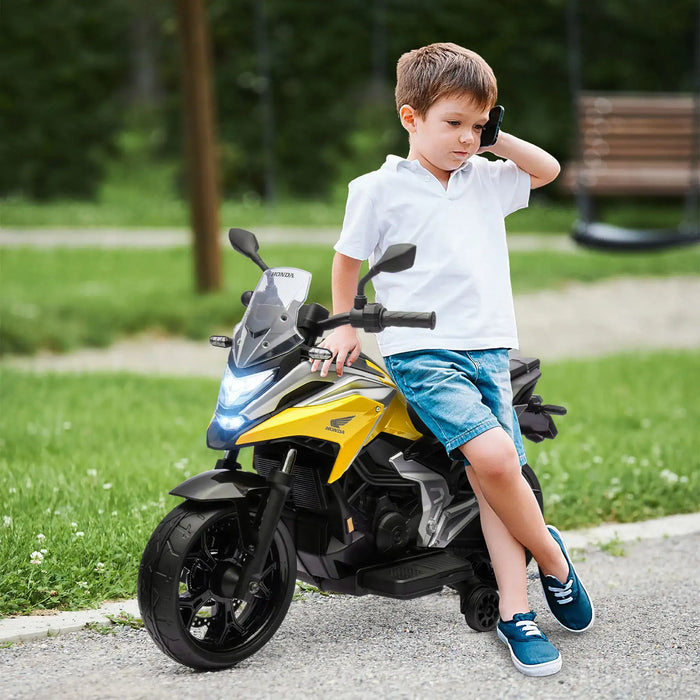 Young boy in white shirt and blue shorts leaning on yellow Honda toy motorcycle with LED lights outdoors