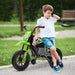 Young boy in white shirt and blue shorts leaning on green electric dirt bike with training wheels outdoors