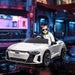 Child in sunglasses driving white Audi RS e-tron GT kids ride-on car with city lights background
