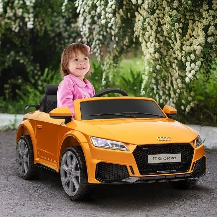 Smiling child in yellow 12V Audi TT RS kids ride-on car outdoors with headlights on