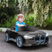 Child driving black BMW i4 kids electric ride-on car outdoors, flowers in background