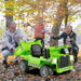 Child playing with green electric ride-on dump truck outdoors with smiling adults in autumn leaves