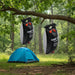 Two 20L solar camp shower bags hanging from a tree branch near a blue tent in a forest.