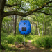 Blue solar camping shower bag hanging from tree in forest with outdoor chairs and table
