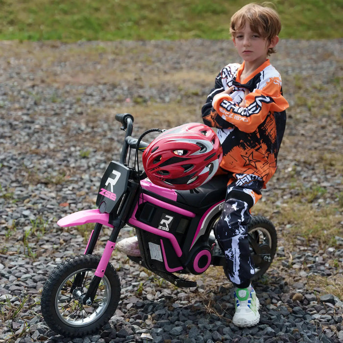 Child in motocross gear next to pink 24V kids electric dirt bike with helmet outdoors