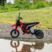 Red 24V kids electric dirt bike on gravel with grass background and water reflection