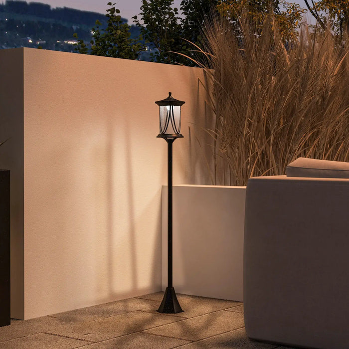 Black outdoor garden lamp post illuminating patio next to beige wall and dried ornamental grass at dusk