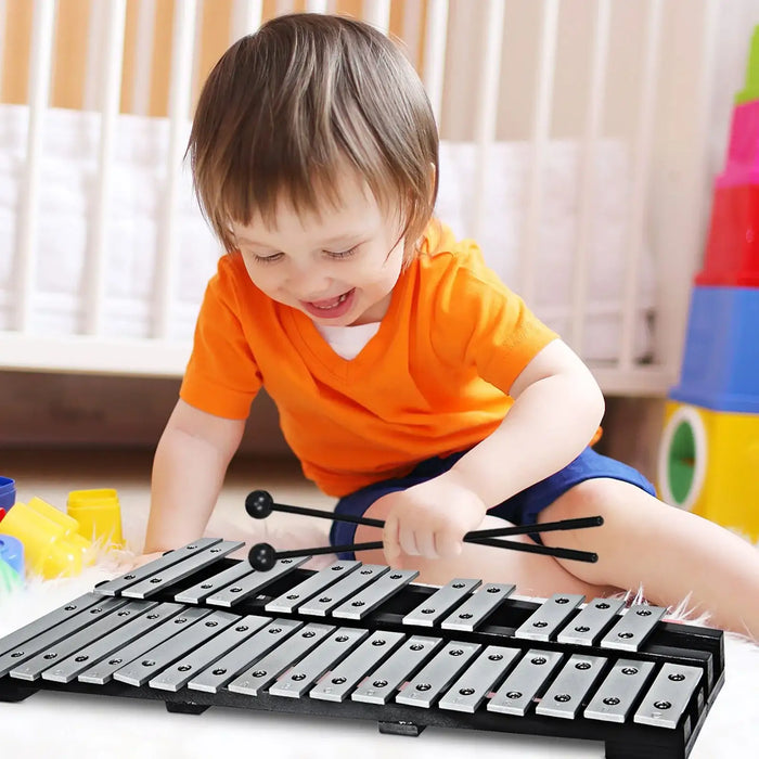 Young child in orange shirt playing silver metal xylophone with black mallets indoors