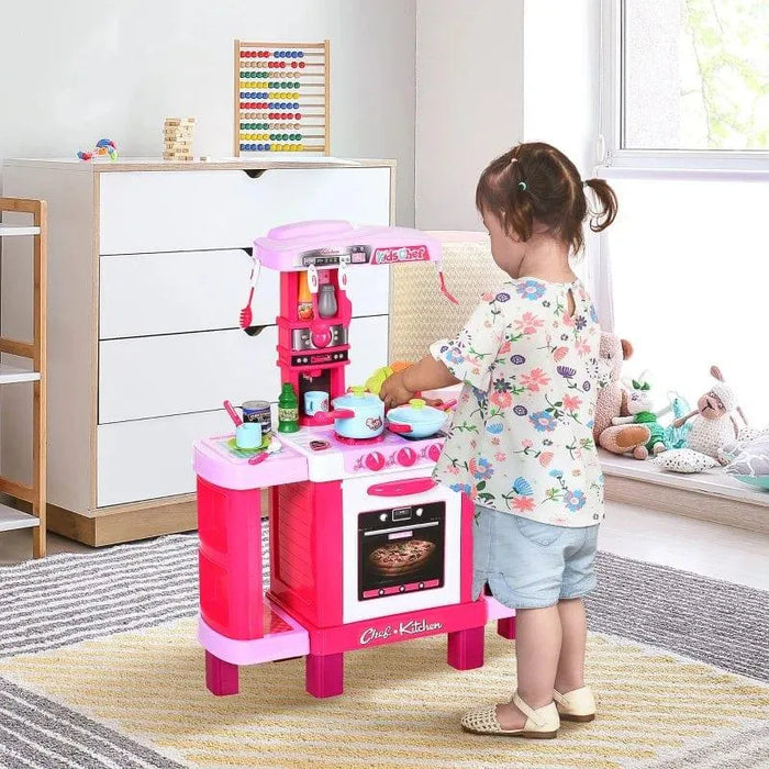 Toddler playing with pink kids kitchen playset with oven, sink, and accessories in a bright room