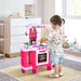 Toddler playing with pink kids kitchen playset with oven, sink, and accessories in a bright room