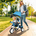 Woman pushing a child in a blue and white tricycle through a park.