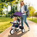Woman pushing a child in a purple and white tricycle through a park.