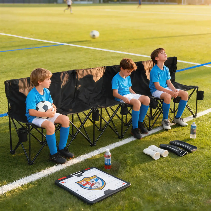 Three boys in blue soccer uniforms sit on a 6-seater folding sports bench with cup holders on a grass field, with soccer gear and water bottles in front.