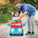 Smiling child riding a red and blue toy car with headlights on, guided by a man outdoors
