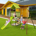 Four children playing on green slide, swings, and sandbox swing set in backyard with yellow house