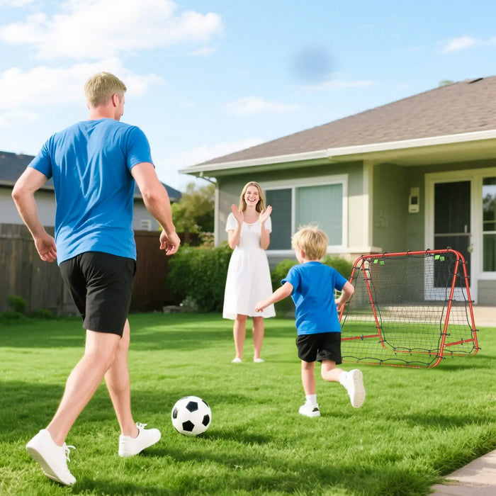 Father and son playing soccer on grass lawn with backyard soccer goal and mother clapping