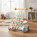 Wooden toy cake with wheels and a handle on a wooden floor, surrounded by books and toys.