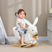 Child sitting on a white rabbit-shaped rocking toy in a room with neutral walls and a lamp.