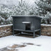 Grey insulated outdoor feral cat house on snow-covered patio with frosted pine trees and stone wall