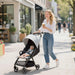Woman pushing a stroller with a baby and holding a coffee, walking on a sidewalk.