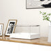 Modern transparent acrylic display case with white base on wooden surface near framed photos and plant