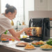 Person using a black air fryer in a kitchen with various food items on the counter.