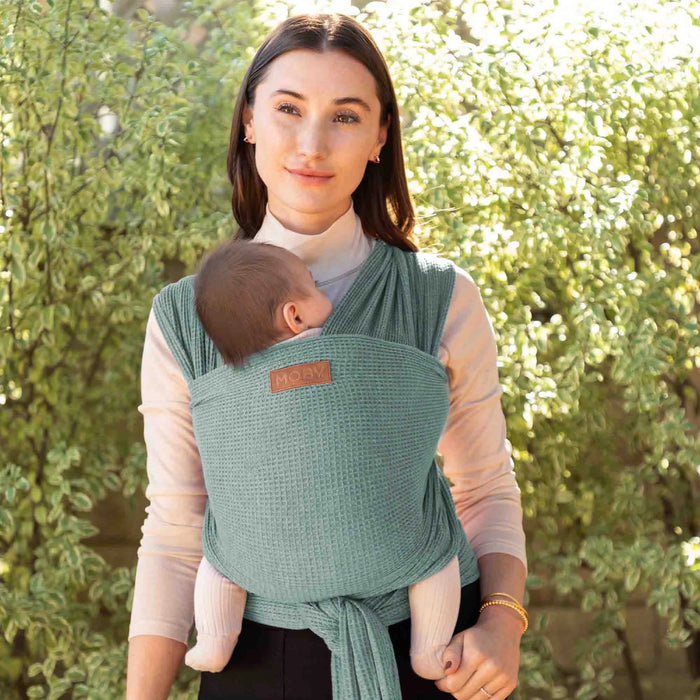 Woman holding a baby in a green baby carrier against a blurred green outdoor background