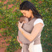 Woman holding a baby in a brown woven wrap against a green leafy background