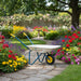 Children's wheelbarrow in a garden setting with colorful flowers and heart-shaped flags on a wooden fence.