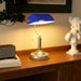 Blue desk lamp on a wooden surface with a plant and sculpture in the background