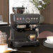Black espresso machine on a table with coffee cups and beans, in a home setting.