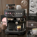 Espresso machine with a glass of coffee on a wooden surface, surrounded by coffee-making accessories.