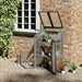 Grey 3-tier wooden mini greenhouse with potted plants on shelves by brick wall