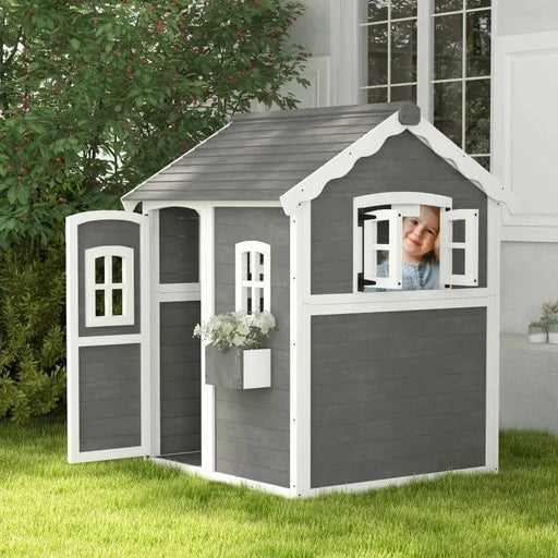 Grey wooden cottage-style playhouse with white trim, open door, flower box, and a child at the window.