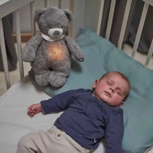 Baby sleeping in crib next to grey teddy bear night light and sound soother