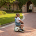 Child riding a toy horse on a brick path with green grass and trees in the background