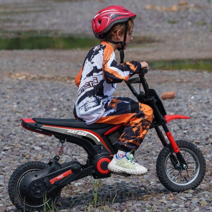 A child wearing a red helmet and colorful motocross gear rides an Electric Dirt Bike with Twist Grip Throttle, Music, Horn and 12" Pneumatic Tyres 24V in Red on a rocky terrain. The 24V motor powers the black and red HOMCOM kids' motorbike as the child appears focused while enjoying the activity. The surrounding area is outdoors with gravel and sparse vegetation.