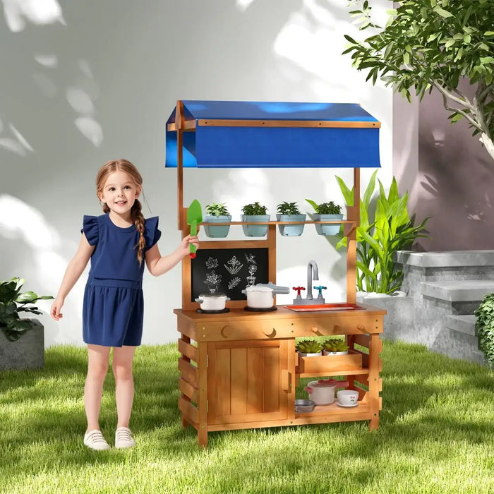 Young girl in blue dress playing with wooden outdoor play kitchen with blue canopy and toy pots