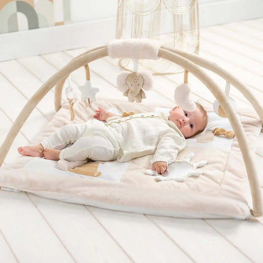 Baby lying on a white play mat with a baby arch and hanging toys in a bright room.