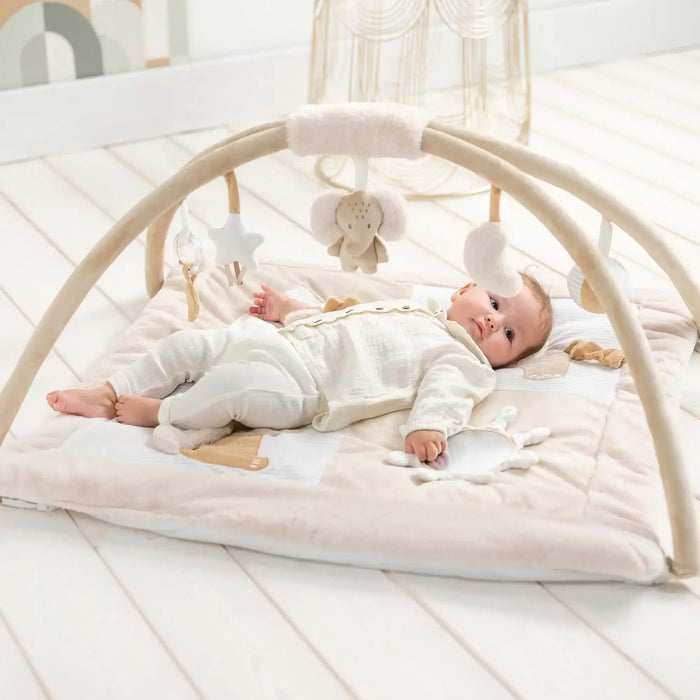 Baby lying on a white play mat with a baby arch and hanging toys in a bright room.