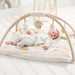 Baby lying on a white play mat with a baby arch and hanging toys in a bright room.
