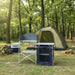 Foldable camping kitchen unit with windscreen and storage set up outdoors by a tent