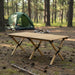 Folding camping table with natural wood effect set in a forest campsite near a green tent.