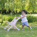 Toddler outdoors pushing pink folding wooden doll stroller on grass under leafy tree