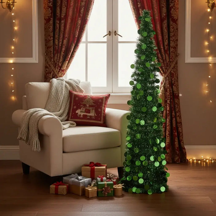 Small green sequin Christmas tree next to armchair with knitted blanket and red pillow, gifts below