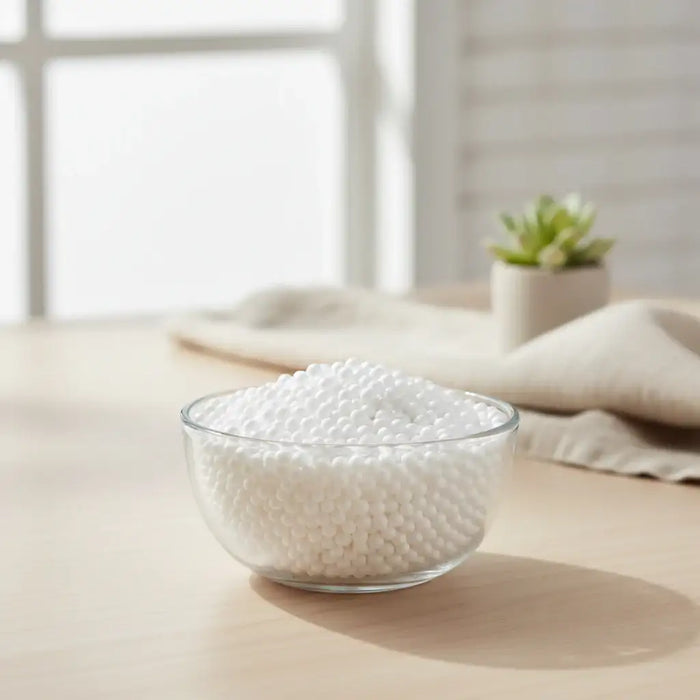 Clear glass bowl filled with white plastic foam beads on a wooden surface with a cloth and small plant in the background