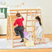 Children playing on a wooden climbing frame in a room with a white wall and wooden floor.