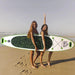 Two women holding inflatable paddleboard on sandy beach, ocean waves in background