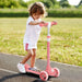 Toddler girl riding pink three-wheeled scooter on paved path in sunny outdoor park